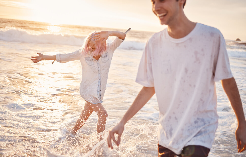 Couple at beach, splash in water with ocean, travel and freedom outdoor, love and care in relationship with youth. Cafe free at sunset, nature and sea waves with young people on holiday in Hawaii